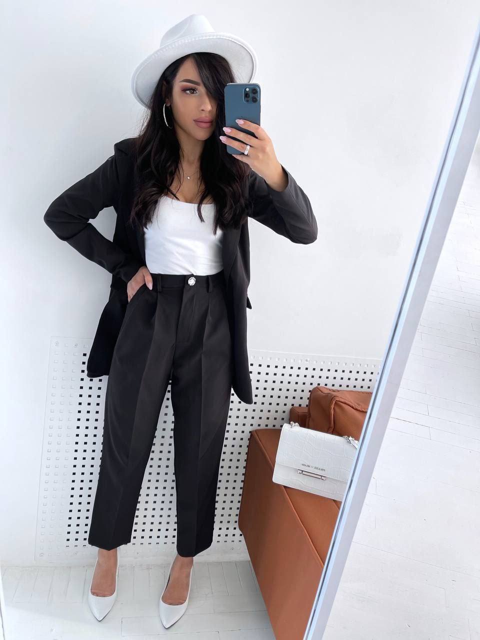 Woman taking a mirror selfie wearing a black blazer, white top, and pants with a white hat.