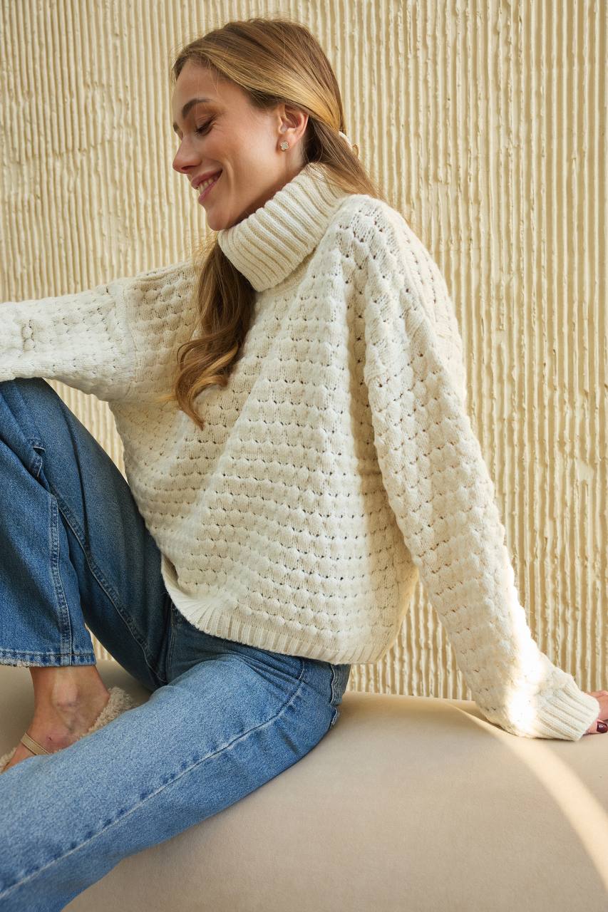 Woman wearing a white chunky knit sweater and blue jeans sitting on a beige chair.
