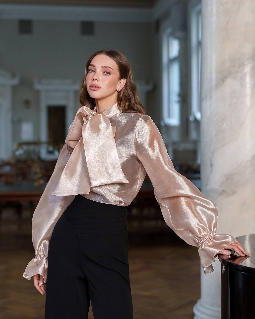 Woman wearing a beige satin blouse and black pants in an elegant interior setting.