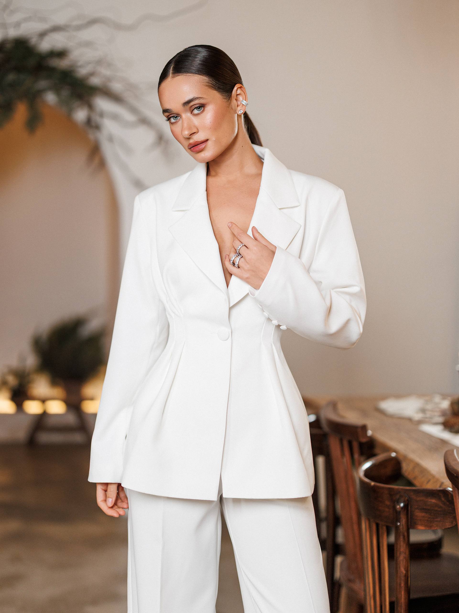 Woman in a white suit standing in an elegant indoor setting with wooden furniture and soft lighting.