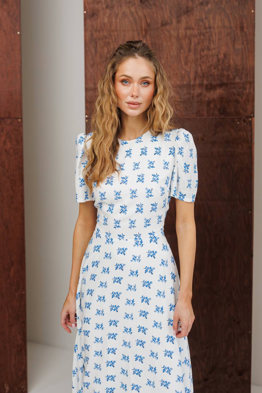 Woman wearing a white dress with blue floral pattern against a wooden wall.