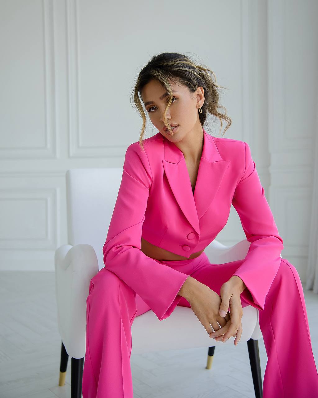 Woman in a bright pink suit sitting on a white chair against a white wall.