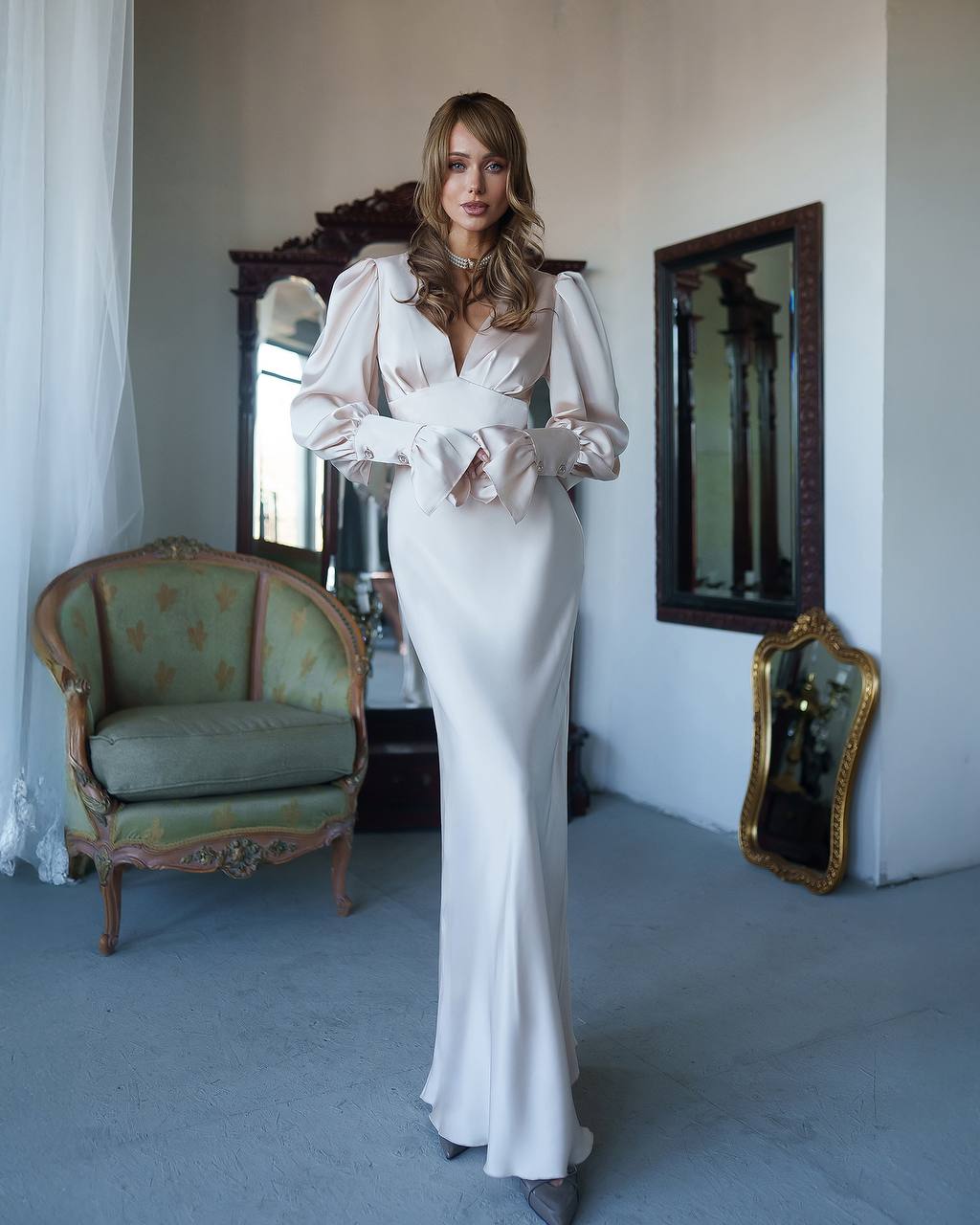 Woman in a white evening gown standing in an elegant room with mirrors and a chair.