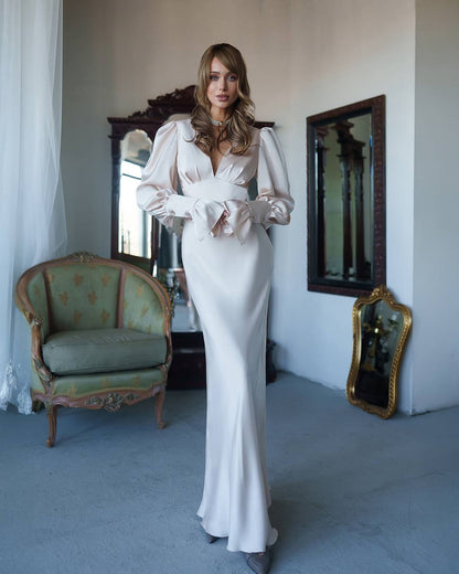 Woman in a white evening gown standing in an elegant room with mirrors and a chair.
