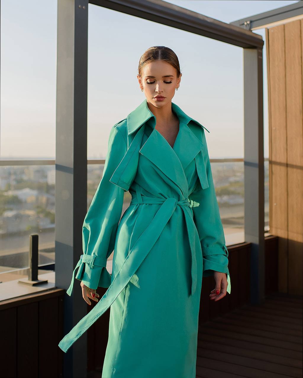 Woman wearing a teal trench coat standing on a rooftop with a cityscape view.