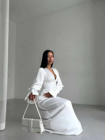 Woman in a white outfit sitting on a white stool against a plain background