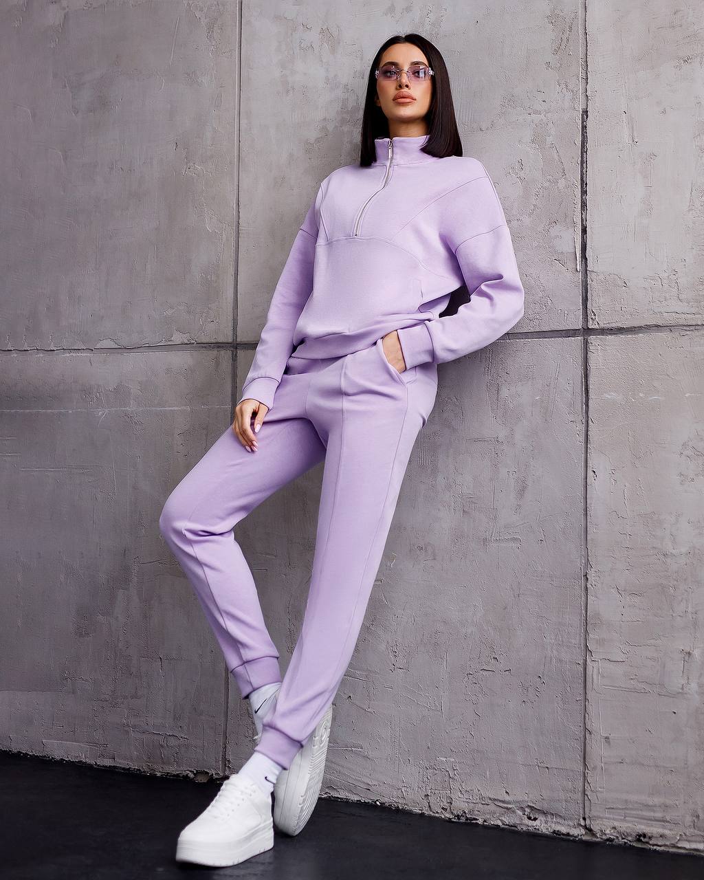 Woman in a lavender tracksuit leaning against a gray concrete wall.