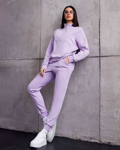 Woman in a lavender tracksuit leaning against a gray concrete wall.