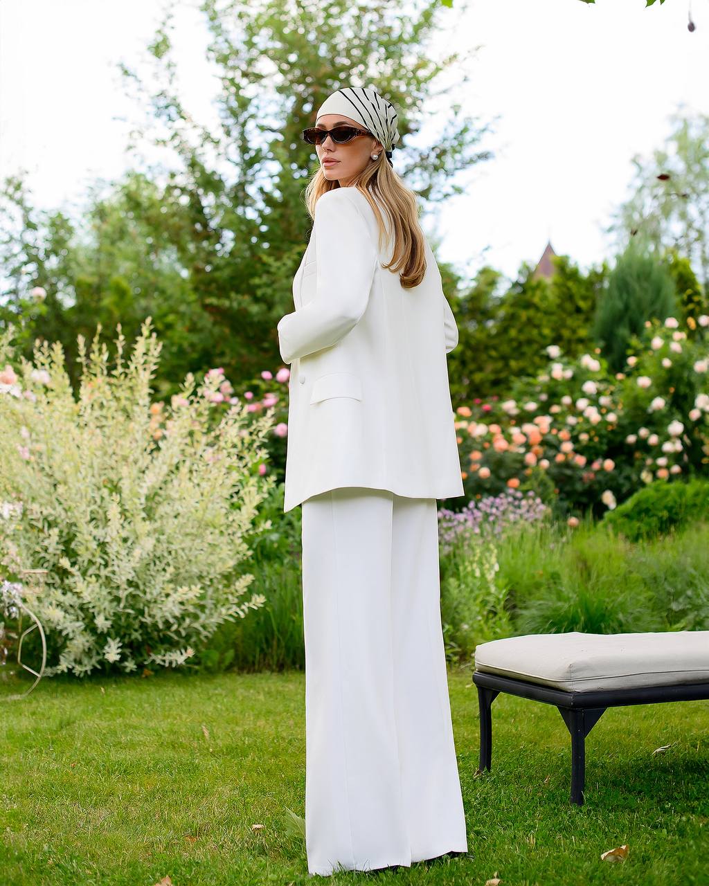 Woman in a white outfit standing in a garden with flowers and a bench in the background