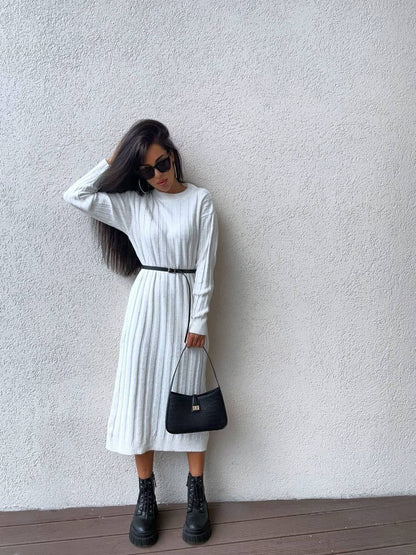 Woman wearing a white dress with a black belt, holding a black handbag against a plain wall.