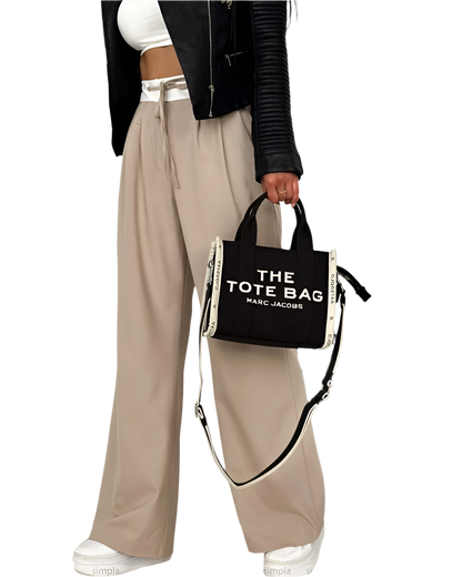 Person holding a black tote bag with 'The Tote Bag' text, wearing beige pants and a black jacket.