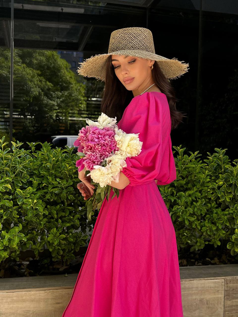 Woman in a bright pink dress and straw hat holding flowers outdoors.