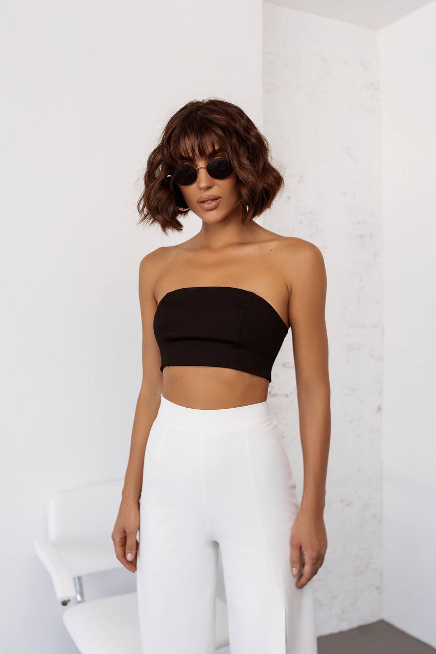 Woman wearing a black strapless top and white pants against a white brick wall.