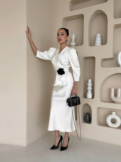 Woman in a white outfit with black accessories standing in a room with decorative shelves.