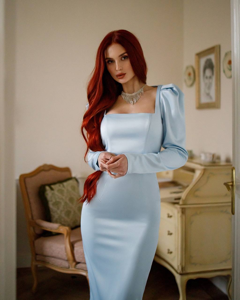 Woman in a light blue dress with long red hair in an elegant room.