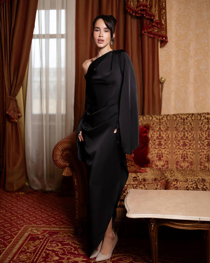 A woman stands in an ornate room wearing a floor-length black one-shoulder gown with draped fabric and a high slit, paired with light-colored heels.