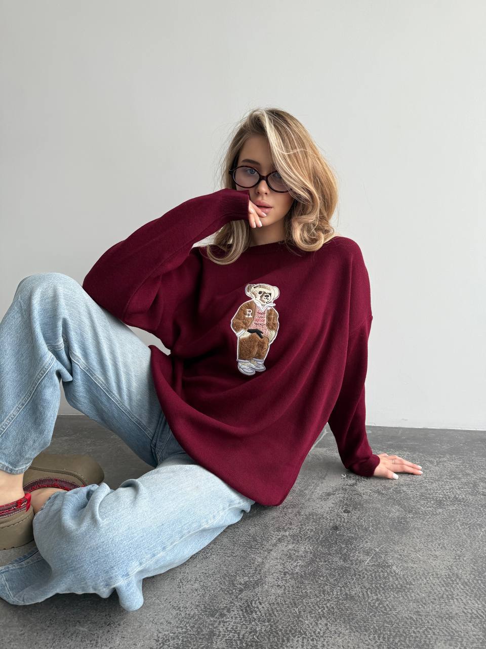 Person wearing a burgundy sweater with a bear graphic, sitting on the floor against a plain background