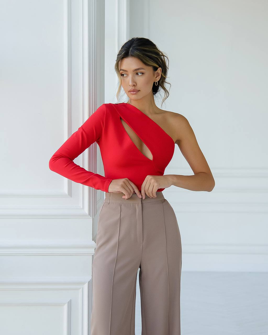 Woman wearing a red one-shoulder top and beige pants against a white wall.
