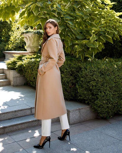 Woman in a beige trench coat standing outdoors with greenery in the background