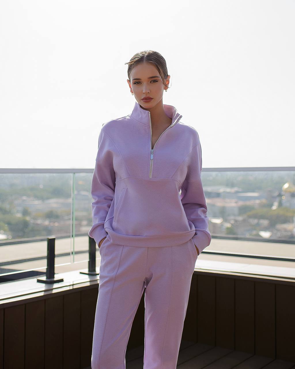 Person wearing a light purple tracksuit standing on a balcony with a cityscape view.