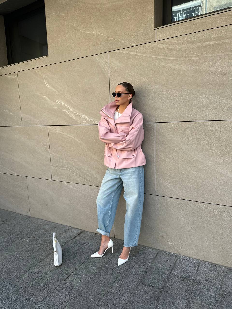 Woman in a pink jacket and blue jeans standing against a beige wall.