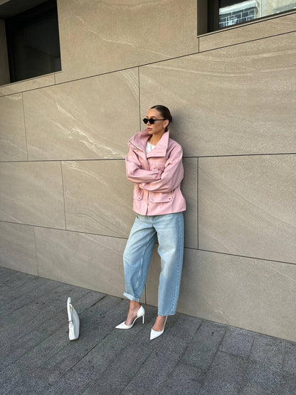 Woman in a pink jacket and blue jeans standing against a beige wall.