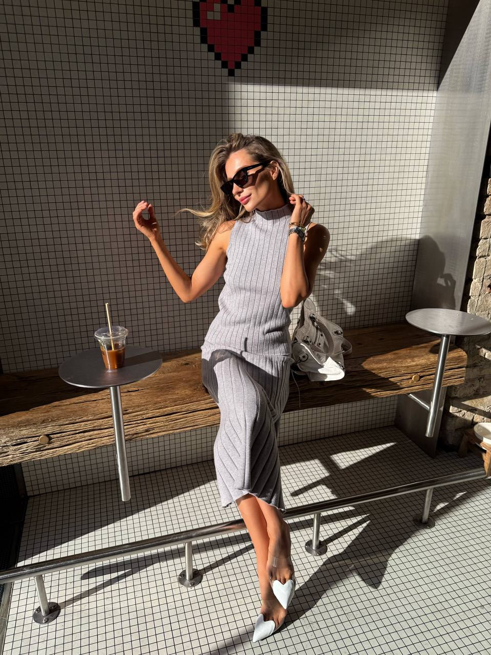 Woman sitting on a bench with a drink, wearing sunglasses and a gray outfit, in an outdoor setting.