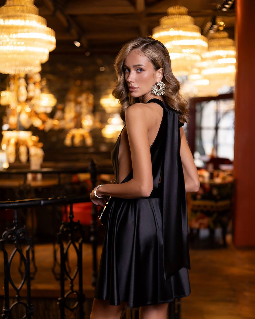 A woman wearing a black satin sleeveless dress with a draped cape-style back, standing in a luxurious indoor setting with glowing chandeliers.