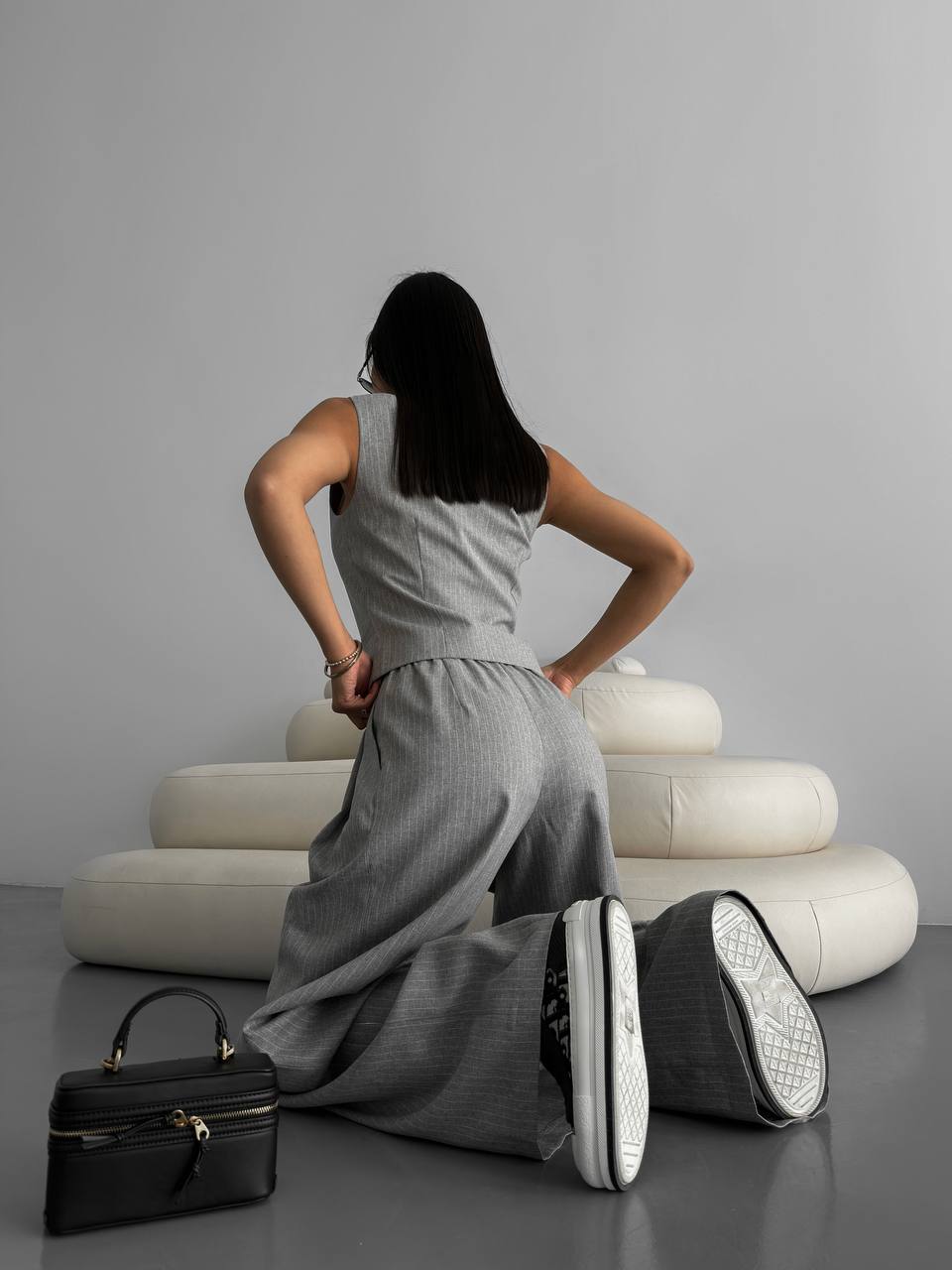 Woman in a gray dress sitting on white cushions with a black handbag beside her.