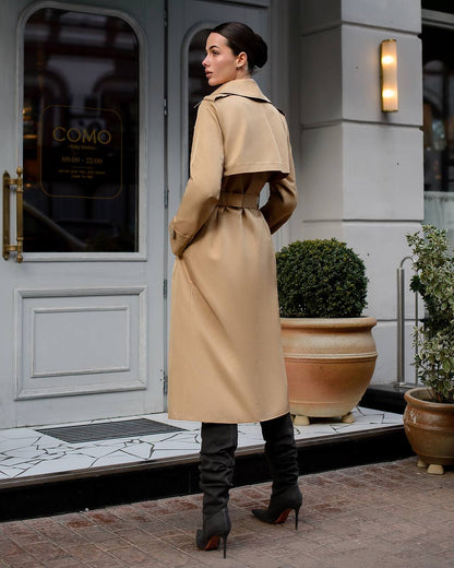 Woman in a beige trench coat standing in front of a building entrance.