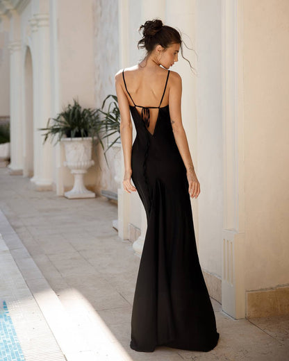 Woman in a black dress standing in a sunlit outdoor setting with columns and plants.