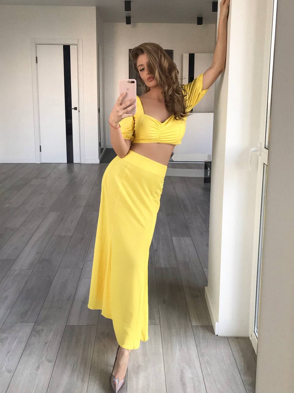 A woman takes a mirror selfie in a bright yellow two-piece outfit consisting of a short-sleeve crop top and a matching long skirt, standing in a modern room with gray wood floors.