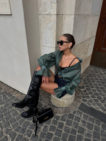 Woman in green shirt and black knee-high boots sitting on a stone bench against a building.