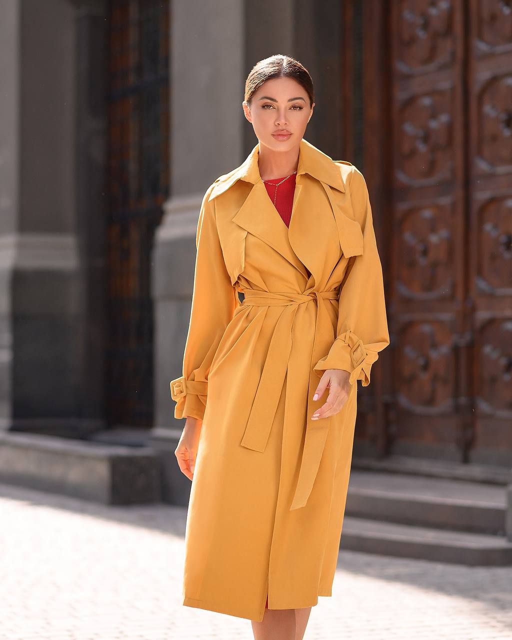 Woman wearing a yellow trench coat standing in front of an ornate door.