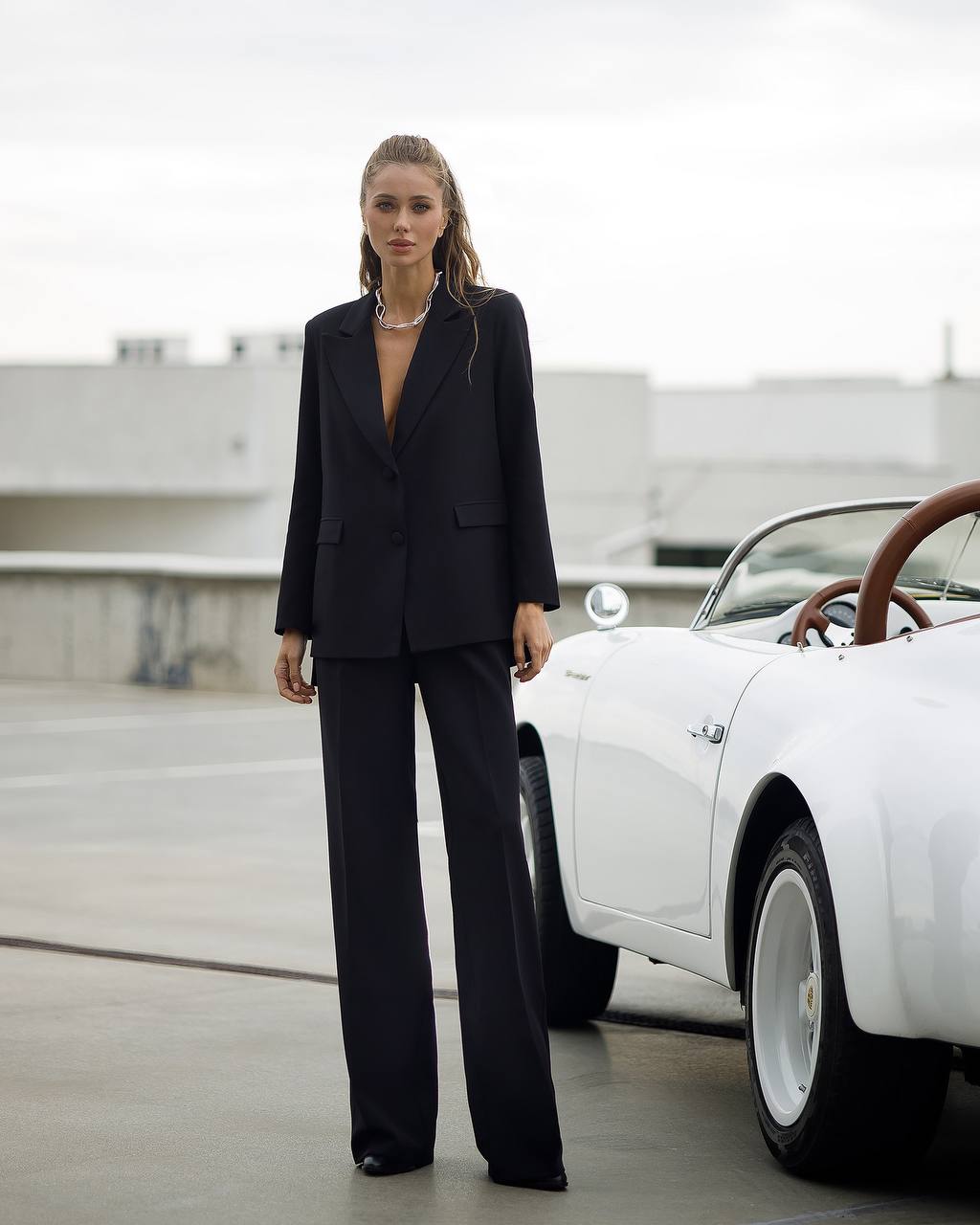 Woman in a black suit standing next to a white vintage car on a rooftop.