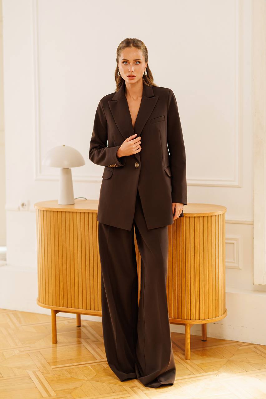 A woman models a dark brown tailored suit consisting of a single-breasted blazer and matching wide-leg trousers, standing in a bright room with light wood furniture.