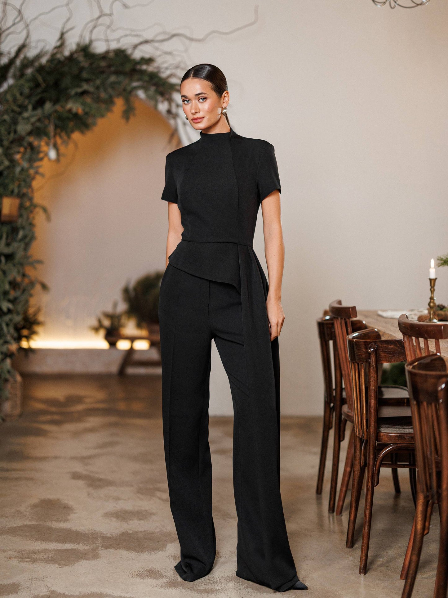 Woman in a black outfit standing in an elegant indoor setting with wooden furniture and decorative plants.