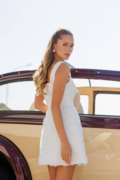 Woman in a white dress standing next to an old-fashioned car with a clear sky background