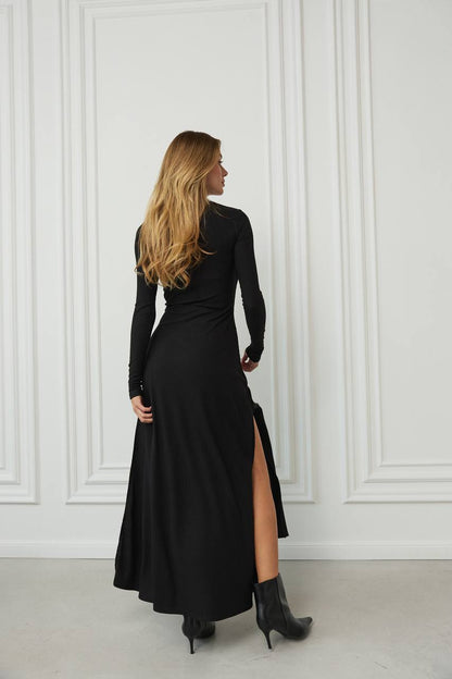 Woman wearing a black dress with a high slit, standing against a white paneled wall.