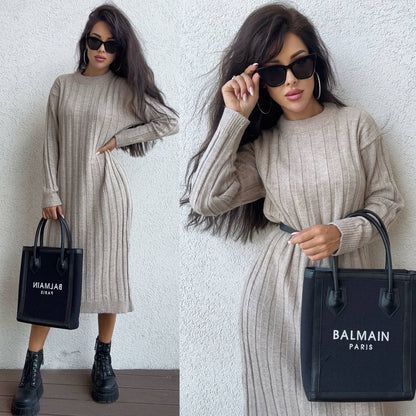 Woman wearing a beige ribbed dress holding a black Balmain handbag against a white wall.