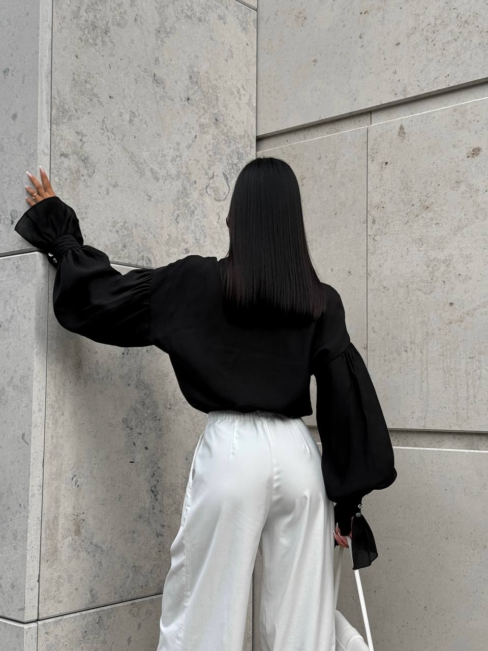 Person wearing a black blouse and white pants against a gray wall