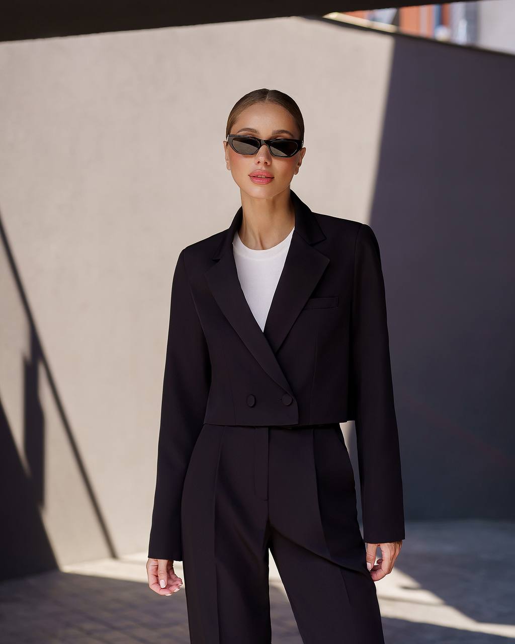 Person wearing a black suit and sunglasses against a neutral wall.