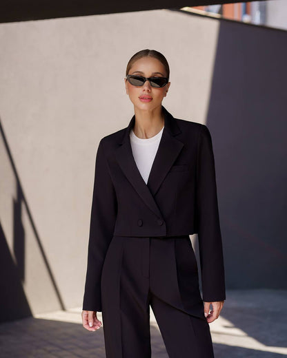 Person wearing a black suit and sunglasses against a neutral wall.