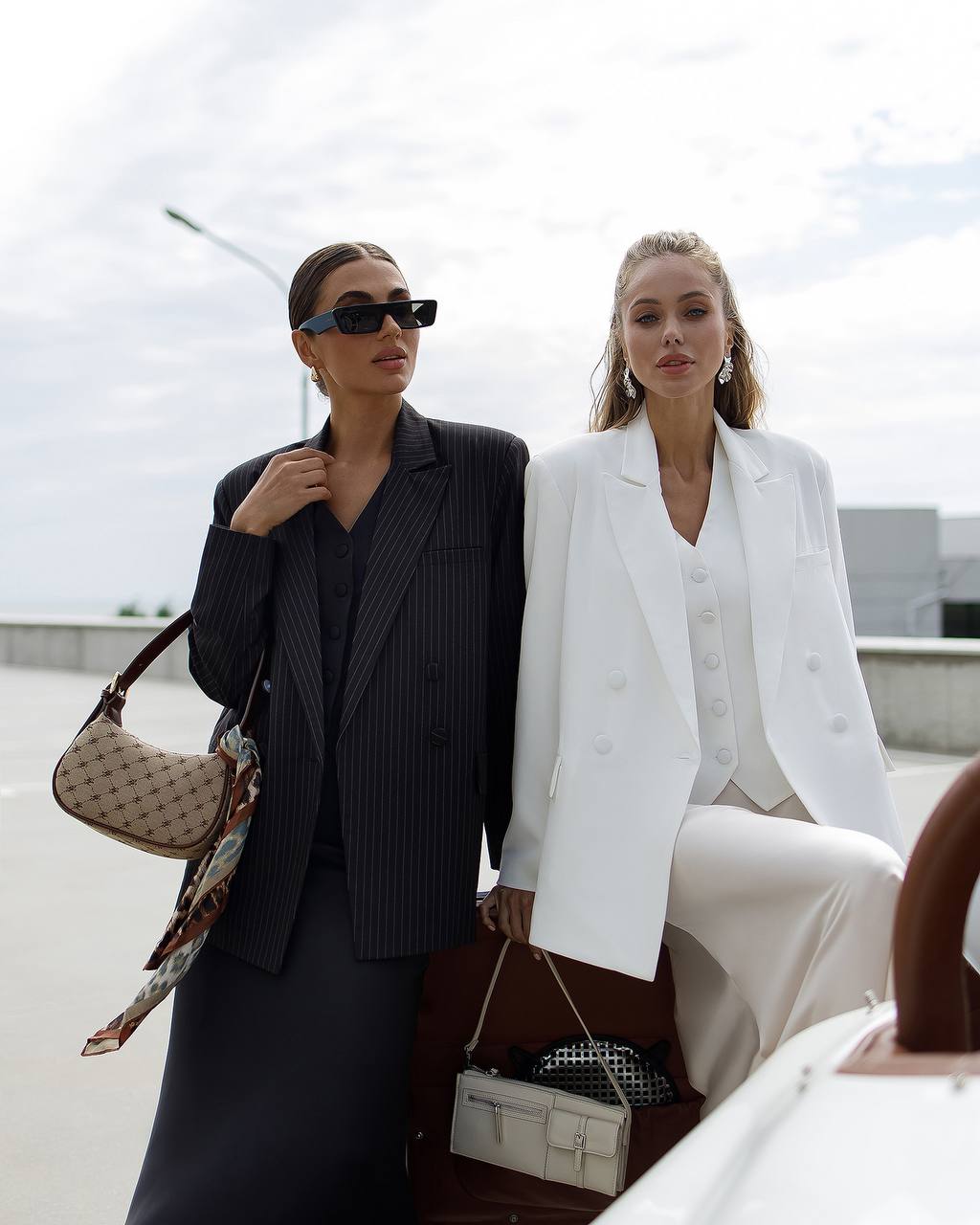 Two women posing outdoors with handbags, one in a black outfit and the other in a white outfit.