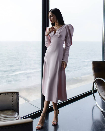 Woman in a light pink dress standing by a large window with a view of water.