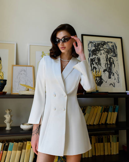 Woman wearing a white blazer dress in an indoor setting with books and artwork.