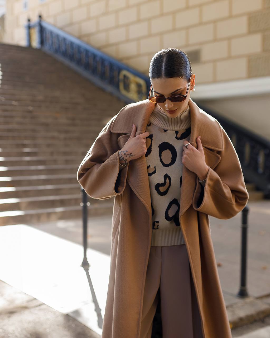 Woman wearing a long beige coat and patterned sweater outdoors.
