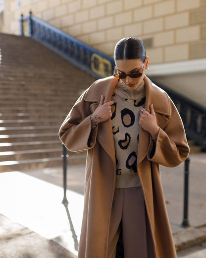 Woman wearing a long beige coat and patterned sweater outdoors.
