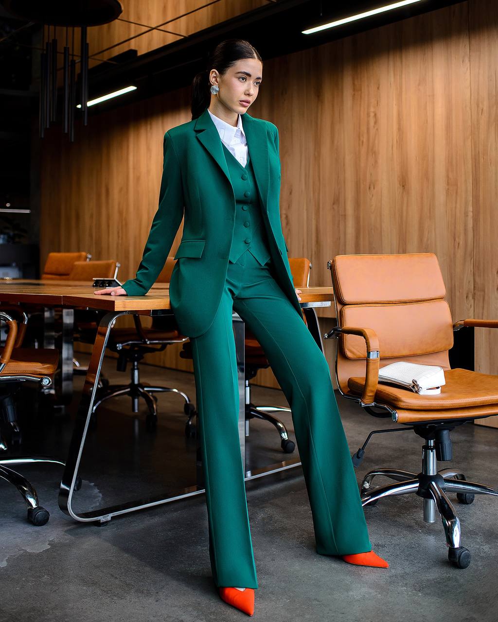Woman in a teal suit standing in an office setting with wooden walls and brown chairs.