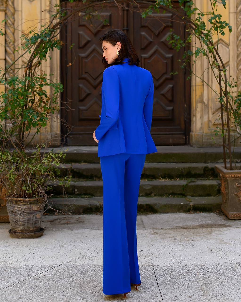 Woman in a blue suit standing in front of a decorative door with plants around.